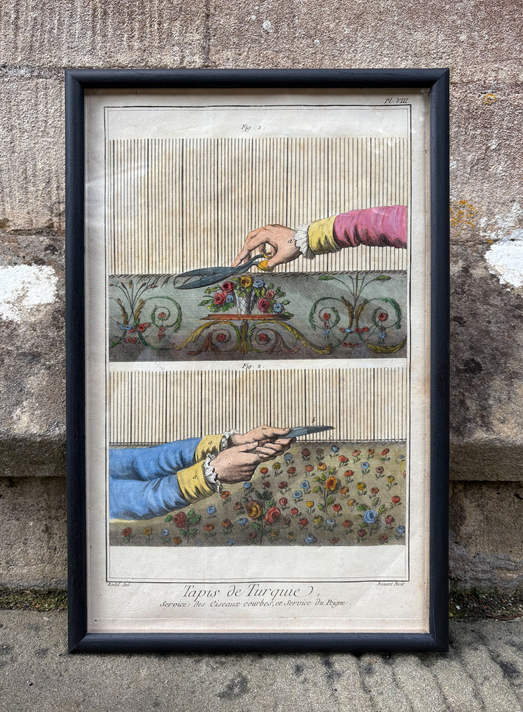 Framed artwork of hands cutting fabric with scissors against a textured stone wall. Antique print of hands trimming a Turkish Carpet 