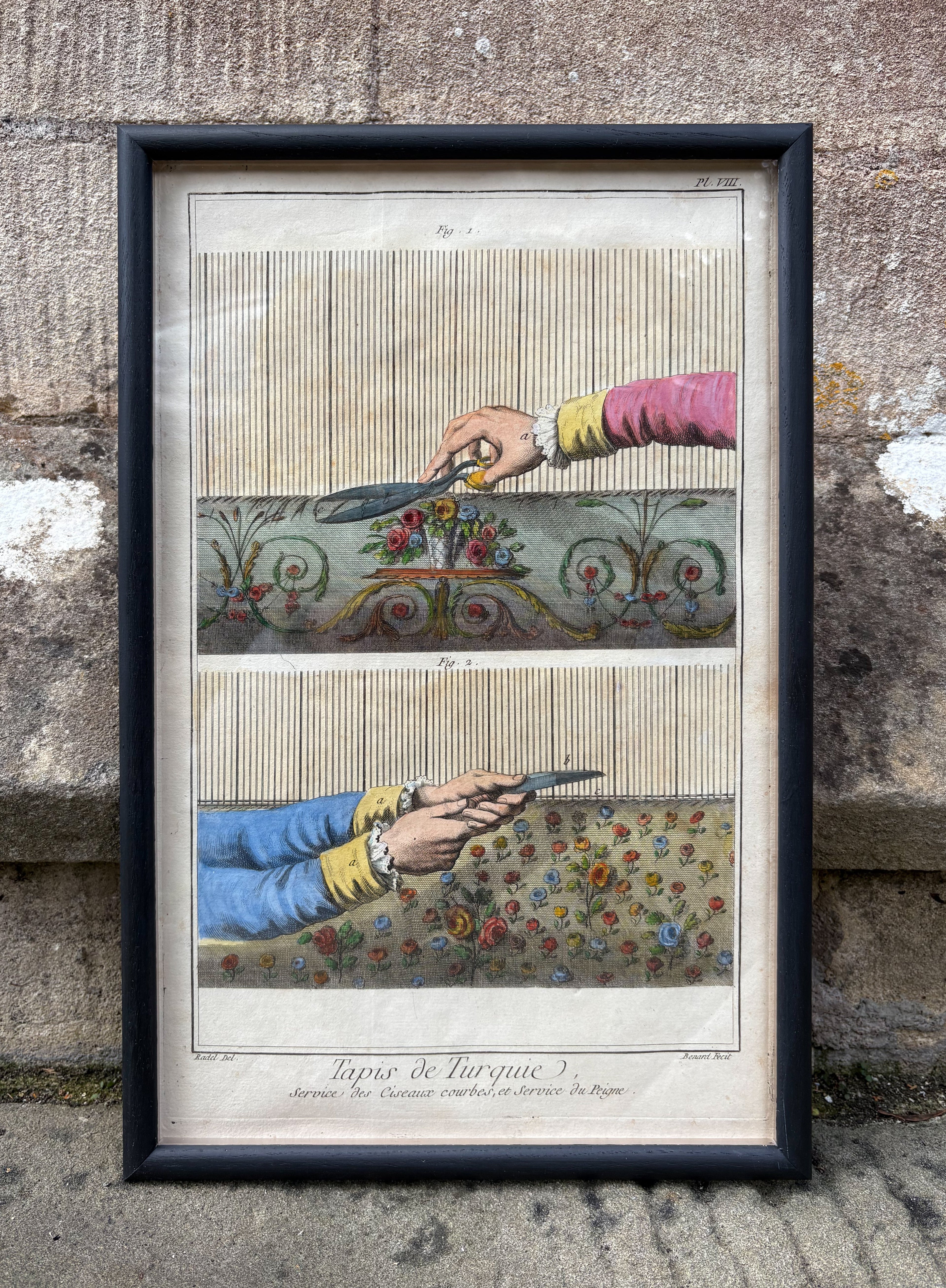Framed artwork of hands cutting fabric with scissors against a textured stone wall. Antique print of hands trimming a Turkish Carpet 